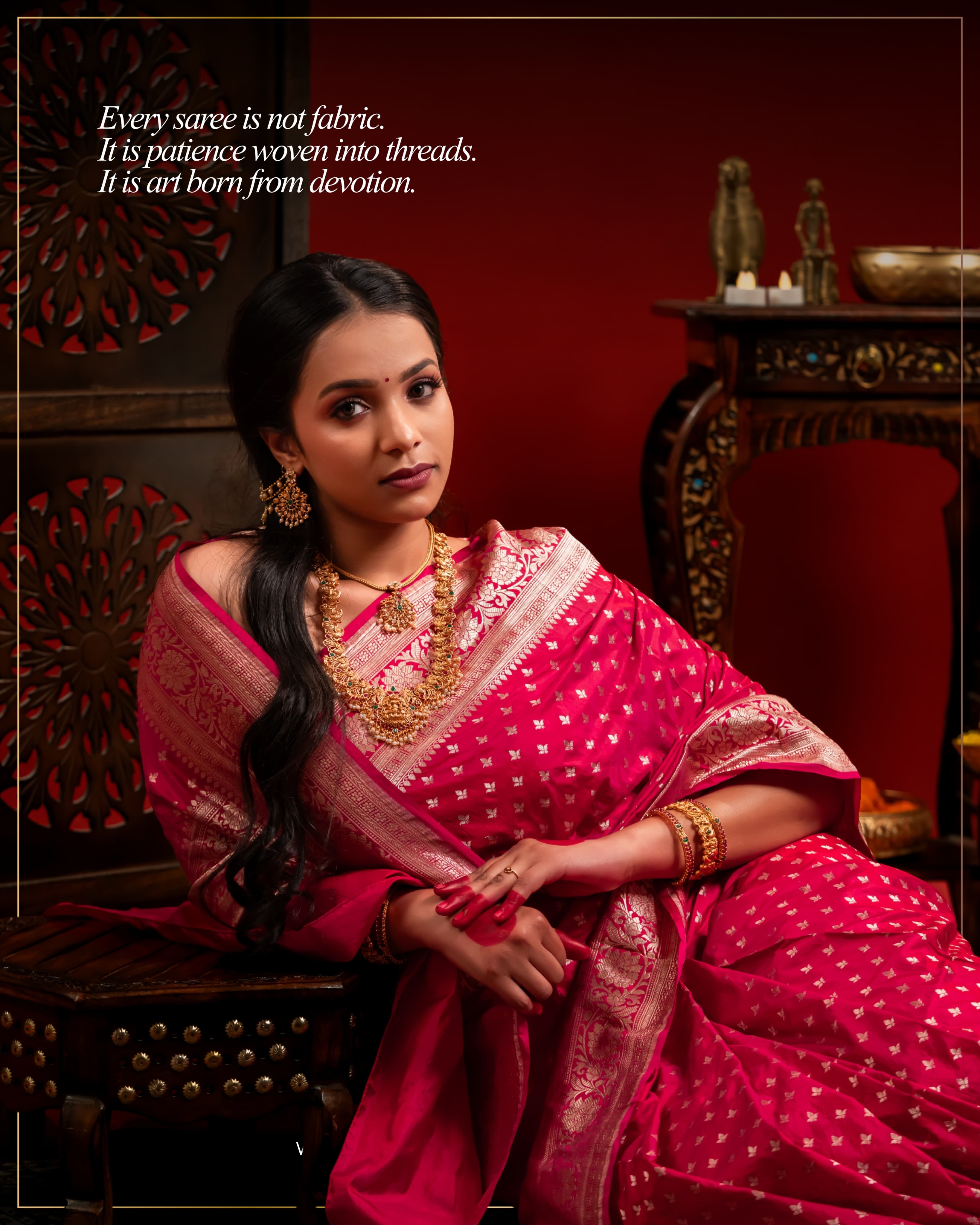 'Every saree is not fabric. It is patience woven into threads. It is art born from devotion' post: a woman in a pink silk saree and temple jewellery seated in a red room with carved wooden furniture