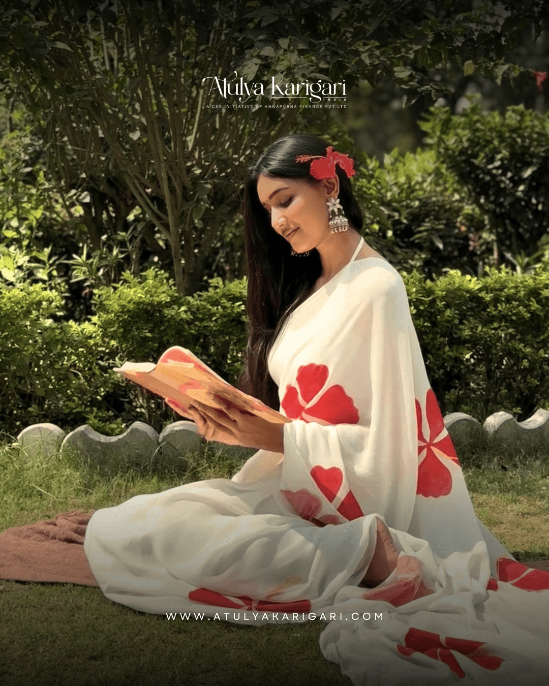 A woman in a white saree with large red floral prints and a red flower in her hair, reading a book on the grass in a garden