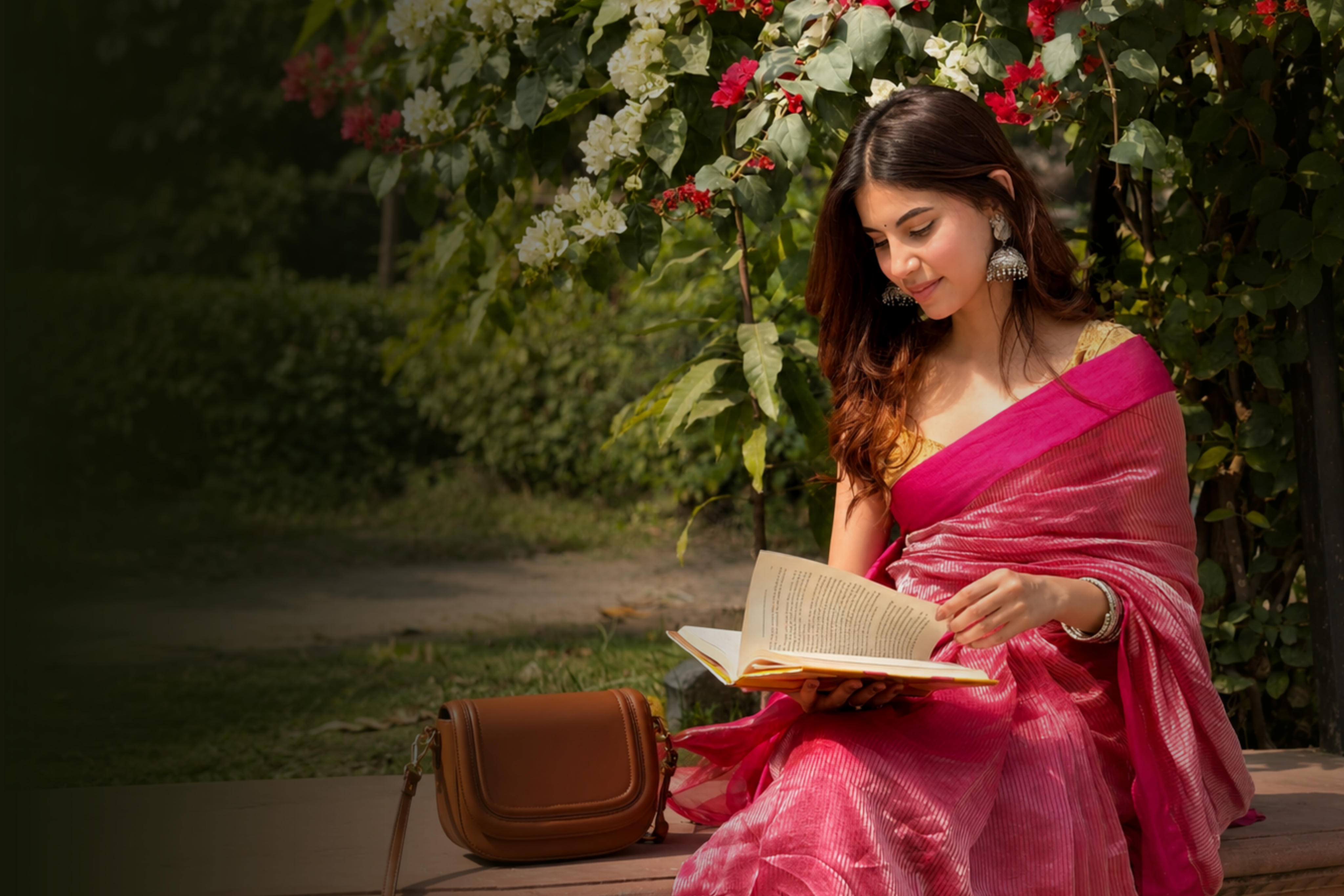 A woman in a pink handloom saree and silver jhumkas reads a book on a garden bench under flowering bougainvillea, a tan leather bag beside her