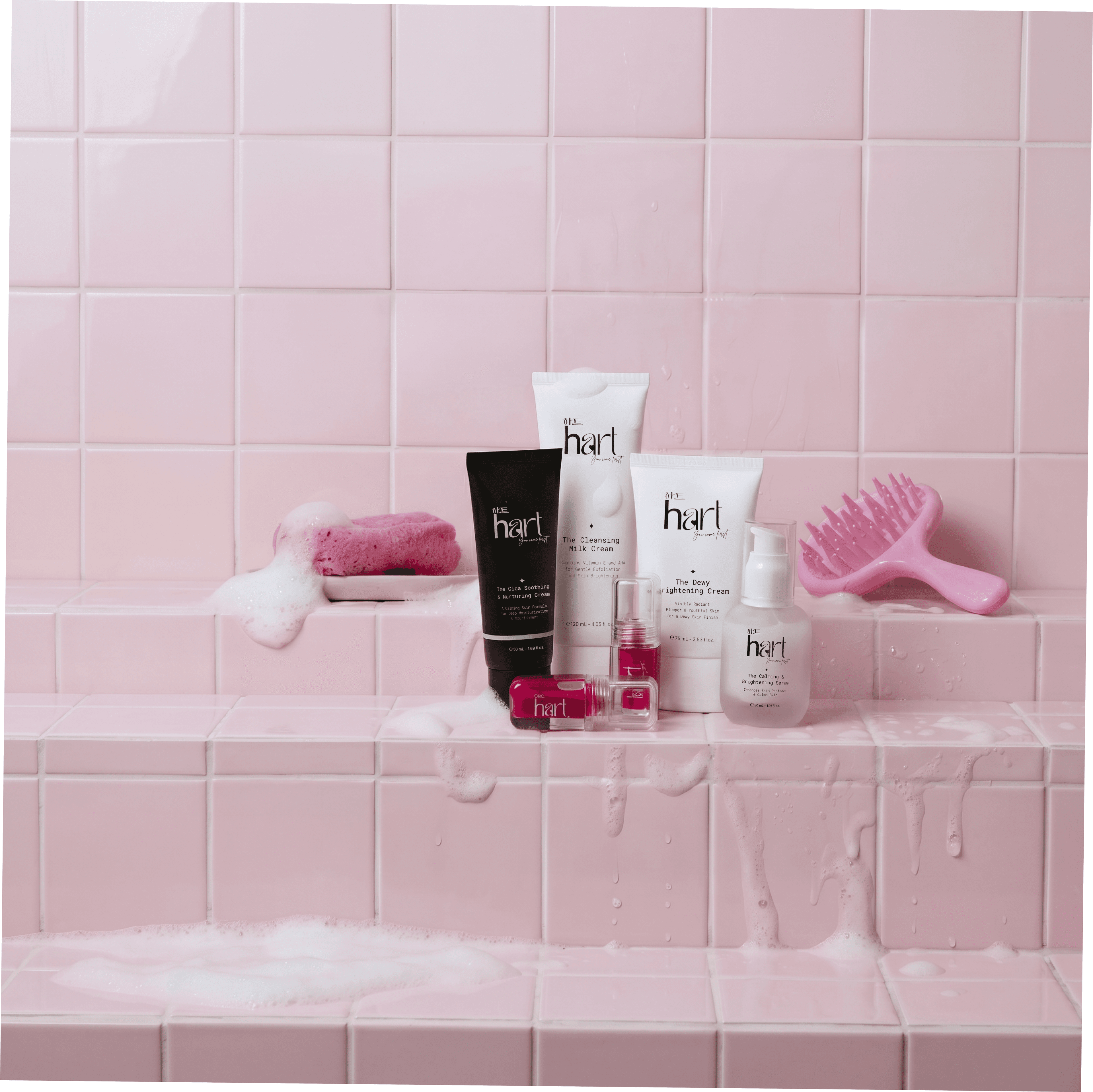 HART skincare on a pink tiled bathroom ledge in soap foam: a black cica cream, white cleansing and brightening creams, a serum bottle and two red lip tints, beside a sponge and a pink scalp brush