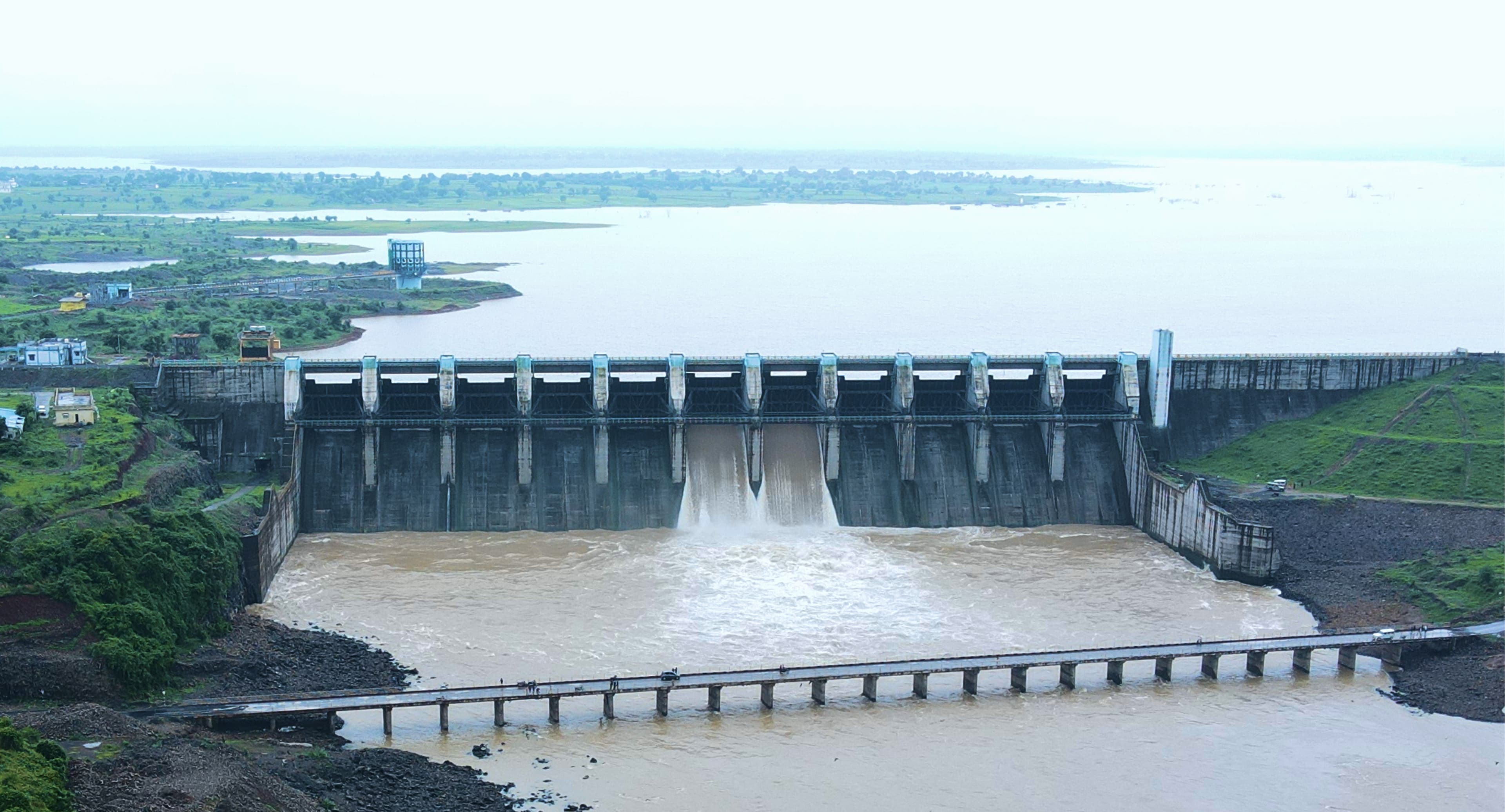Aerial view of a dam with water released through its open gates into the river below, a low bridge crossing in the foreground and the reservoir stretching behind