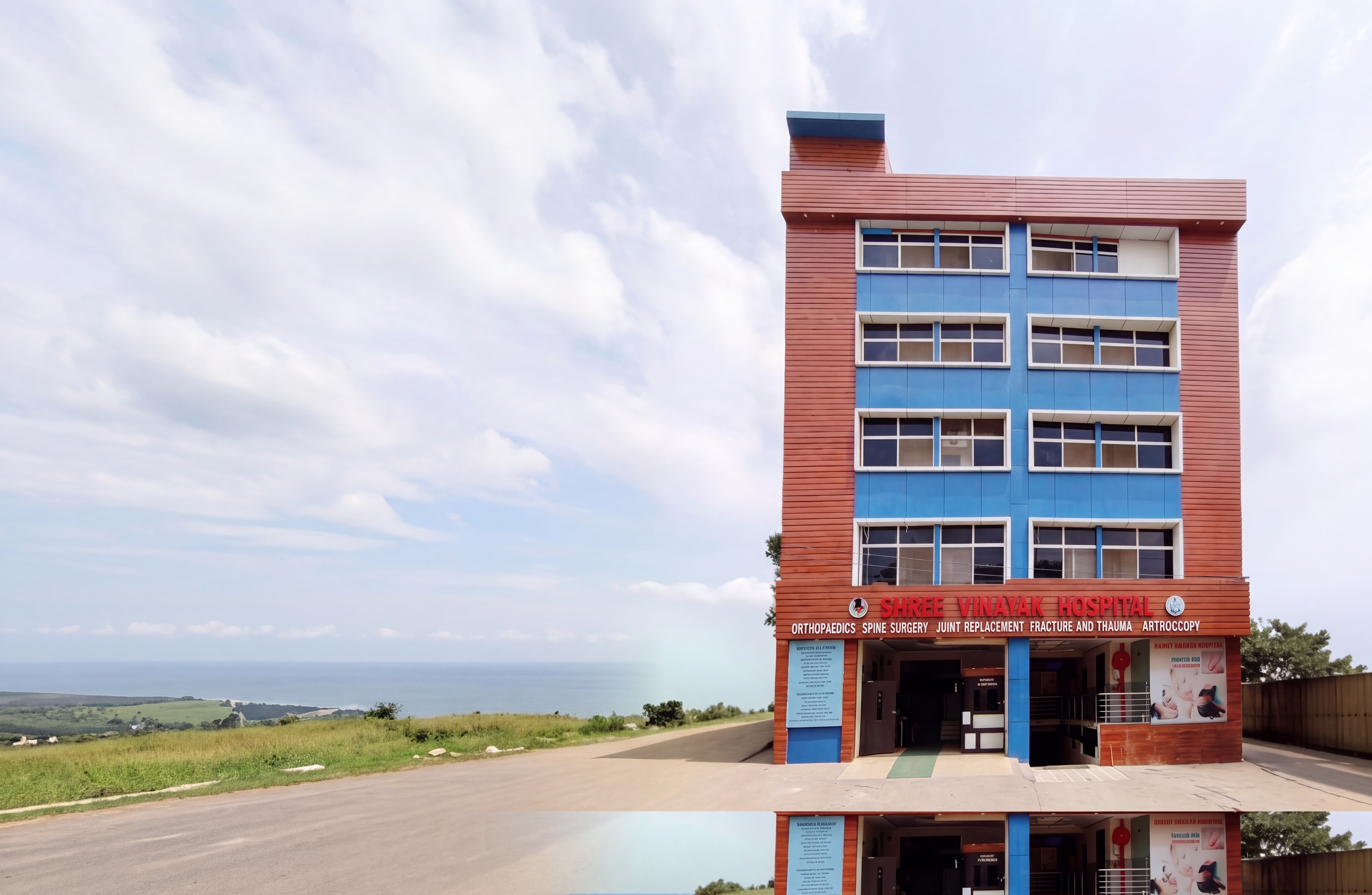 The Shree Vinayak Hospital building, a five-storey block in terracotta cladding and blue panels under the red hospital sign, against a wide cloudy sky